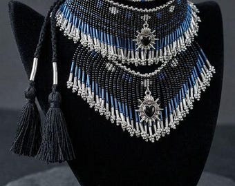 May include: A multi-layered beaded choker necklace featuring black and blue seed beads, silver-toned tube beads, and two silver-toned heart-shaped charms with black centers. The necklace includes long black cord ties finished with black tassels.