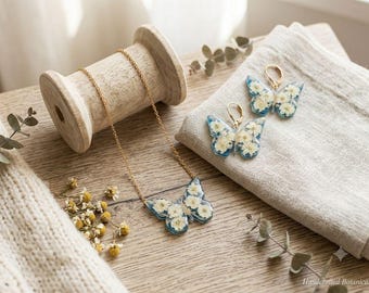 May include: A gold-tone chain necklace and a pair of matching lever-back earrings, each featuring a butterfly-shaped pendant made of clear resin embedded with small white pressed flowers and a blue-tinted base.