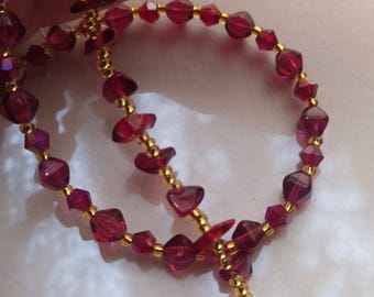 May include: A beaded necklace featuring translucent, deep red, faceted, irregular-shaped beads alternating with small, metallic gold-colored seed beads. The strand is arranged in a circular loop, showcasing the contrast between the red and gold.