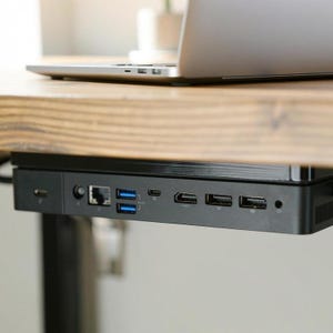 Könnte beinhalten: Eine schwarze Dockingstation mit verschiedenen Anschlüssen, darunter USB, HDMI und Ethernet, ist unter einem Holztisch montiert. Ein silberner Laptop liegt auf dem Tisch. Die Dockingstation bietet Konnektivität und Organisation.