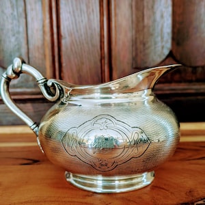 May include: A silver creamer with a curved handle and spout. The body has an embossed design with a central cartouche. The creamer sits on a round base and is displayed on a wooden surface.