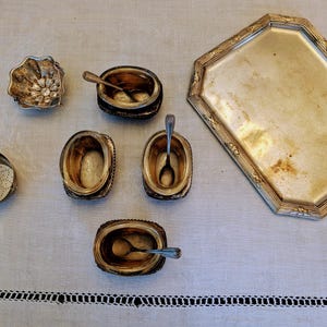 Pode incluir: Uma variedade de peças de servir antigas em prata, incluindo pequenas tigelas com colheres, um coador e uma bandeja decorativa. As peças estão dispostas sobre uma superfície branca, mostrando seus intrincados desenhos e pátina envelhecida.