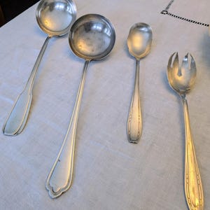 May include: Four pieces of silver serving cutlery are arranged on a white tablecloth. The set includes two ladles, a serving spoon, and a serving fork. The utensils have long, slender handles and rounded or oval bowls.