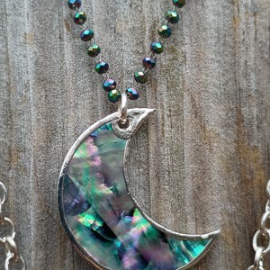 May include: A crescent moon pendant necklace with a silver-tone frame and iridescent abalone shell inlay. The necklace features a beaded chain with iridescent beads. The pendant is approximately 2 inches wide.