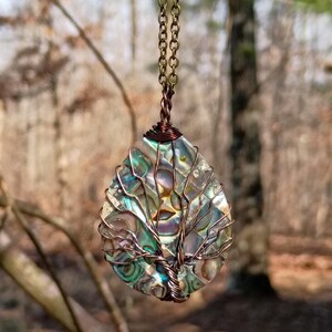 May include: A teardrop-shaped pendant necklace featuring a colorful abalone shell encased in copper wire. The wire is shaped into a tree design. The pendant hangs from a delicate copper chain, creating a nature-inspired accessory.