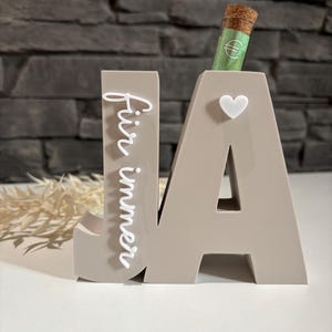 Könnte beinhalten: Eine dekorative beige Holzskulptur mit der Aufschrift "JA" und dem Wort "Für immer" in weißer Schrift auf dem Buchstaben "J". Ein kleines weißes Herz ist am Buchstaben "A" befestigt. Eine kleine Glasampulle mit Korken und einem Euro-Schein steckt oben im "A".