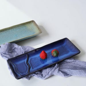 May include: Two rectangular ceramic serving platters. One is a gradient of blue and green, the other is a deep blue with a glossy finish. Two red strawberry-shaped candies rest on the blue platter.