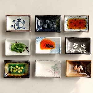 May include: Nine rectangular ceramic dishes with various contents and designs. Some dishes contain food, such as seaweed, blueberries, and peanuts. Others feature floral patterns and abstract designs in shades of blue, brown, and white.