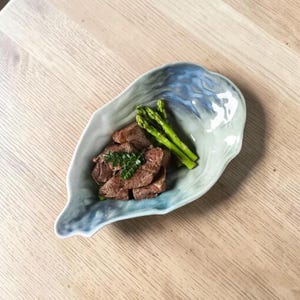 Può includere: Un piatto da portata in ceramica a forma di foglia con smalto blu e bianco. Il piatto contiene carne cotta, punte di asparagi verdi e un rametto di erbe fresche. Il piatto è appoggiato su una superficie di legno chiaro.