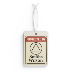 Puede incluir: Ambientador rectangular crema con borde rojo superior con texto "PROTECTED BY". Presenta un triángulo negro dentro de un círculo y el texto "Smith & Wilson". Una cuerda blanca está adjunta.
