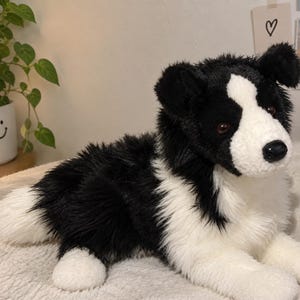 May include: A plush stuffed animal designed to resemble a Border Collie dog. The toy features long, soft black and white fur, with a white blaze running up the center of its face, dark brown eyes, and a black nose. It is depicted in a lying position.