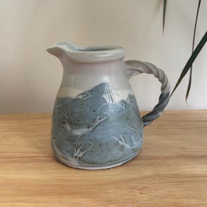 May include: A handmade ceramic jug with a twisted handle. The jug features a light blue and white colour scheme with a mountain design. The jug is sitting on a wooden surface.