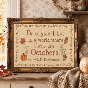 May include: A framed cross-stitch sampler on beige fabric featuring the text "I'm so glad I live in a world where there are Octobers. - L.M. Montgomery" surrounded by a decorative border of embroidered autumn leaves, a pumpkin, and an acorn.