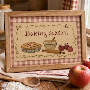 Peut inclure: Un cadre contenant une broderie au point de croix avec le texte Baking season. Le motif représente une tarte, un bol avec une cuillère en bois, un rouleau à pâtisserie et trois pommes rouges, le tout entouré d'une bordure sur un tissu beige.