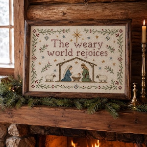 Puede incluir: Un cuadro de punto de cruz con el texto "The weary world rejoices" en letras de color rojo oscuro. El diseño incluye una escena de nacimiento con figuras en un establo, dos ovejas y una estrella dorada, todo rodeado por un borde floral verde y rojo.