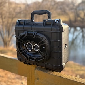 Op de afbeelding: Een zwarte draagbare luidspreker met een handvat en een groot luidsprekerrooster. De luidspreker heeft een robuust, boxy ontwerp en is gemonteerd op een houten hek. De luidspreker heeft twee ronde luidsprekercomponenten.
