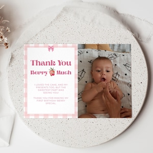 May include: A baby and a pink and white card with the text "Thank You Berry Much" are displayed on a white, speckled surface. The card has a gingham border and a strawberry graphic. The baby is lying on a patterned blanket.