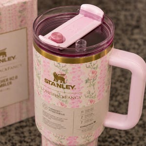 Może przedstawiać: R&oacute;żowy kubek Stanley Loveshackfancy Quencher H2.0 z kwiatowym wzorem. Kubek ma r&oacute;żowy uchwyt i pokrywkę ze złotą opaską. Pudełko obok ma ten sam kwiatowy wz&oacute;r i logo Stanley.