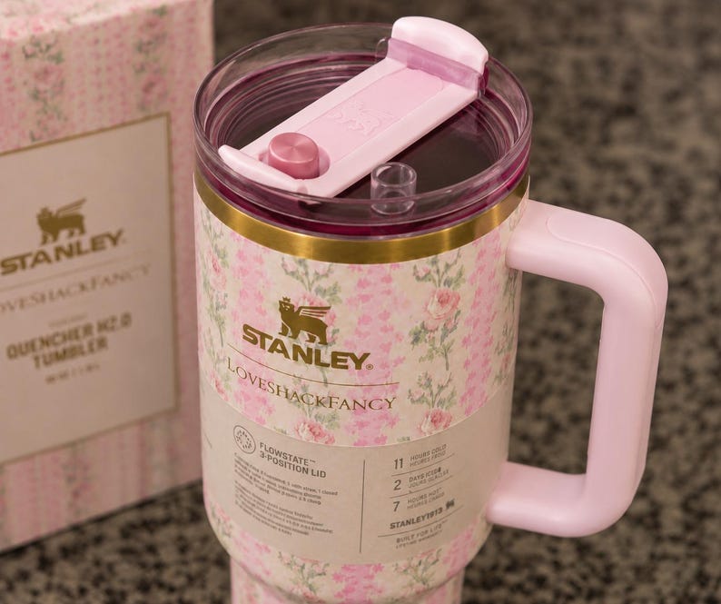 Może przedstawiać: R&oacute;żowy kubek Stanley Loveshackfancy Quencher H2.0 z kwiatowym wzorem. Kubek ma r&oacute;żowy uchwyt i pokrywkę ze złotą opaską. Pudełko obok ma ten sam kwiatowy wz&oacute;r i logo Stanley.