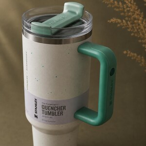 Puede incluir: Un vaso Stanley Quencher blanco y gris moteado con un asa y tapa verde menta. El vaso tiene el texto "THE FLOWSTATE QUENCHER TUMBLER" y "40 OZ 1.18L" impreso. Dise&ntilde;ado para la hidrataci&oacute;n durante todo el d&iacute;a.
