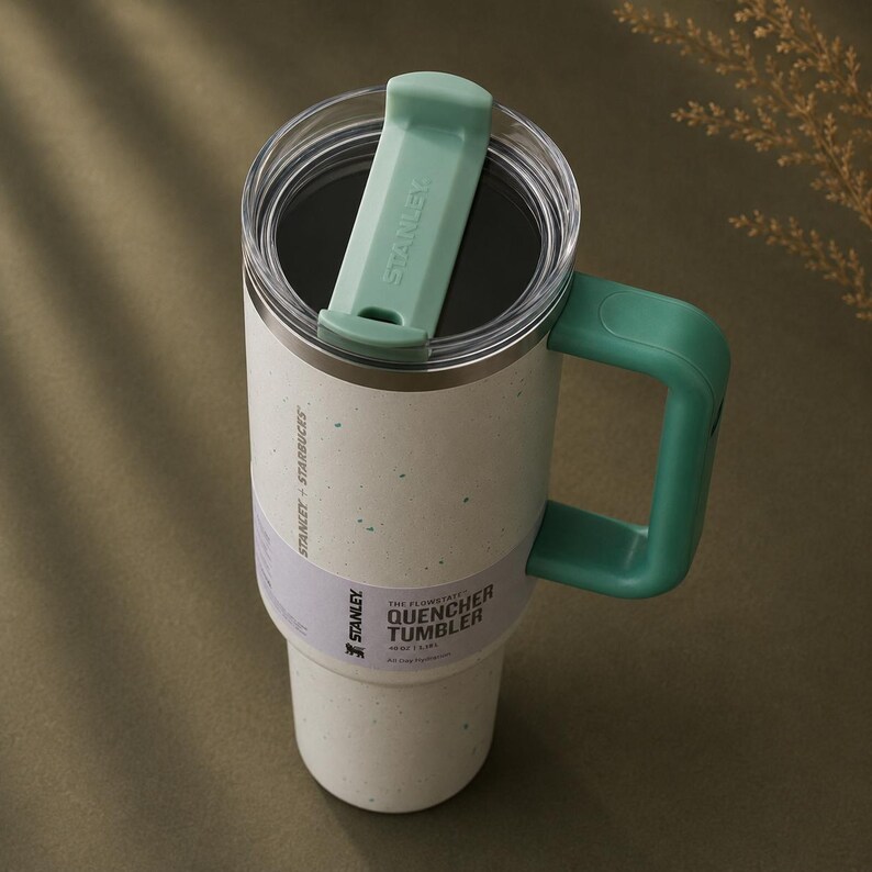 Puede incluir: Un vaso Stanley Quencher blanco y moteado con asa y tapa verdes. El vaso tiene una tapa transparente y una etiqueta que dice "Stanley + Starbucks". El vaso est&aacute; dise&ntilde;ado para mantener las bebidas fr&iacute;as durante horas.