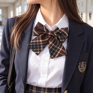 May include: A dark brown plaid bow tie worn over a crisp white collared button-down shirt, layered beneath a navy blue blazer featuring a gold embroidered crest on the chest pocket and a matching plaid skirt visible at the bottom edge.