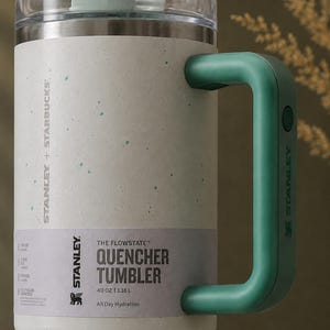 Puede incluir: Un vaso Stanley Quencher moteado blanco y verde azulado con asa y tapa verde azulado. El vaso tiene el texto "STANLEY + STARBUCKS" y "THE FLOWSTATE QUENCHER TUMBLER 1,18 L" impreso en el lateral. Dise&ntilde;ado para la hidrataci&oacute;n durante todo el d&iacute;a.