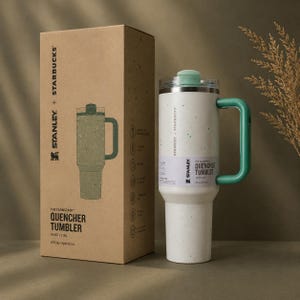 Puede incluir: Un vaso Stanley + Starbucks Quencher salpicado blanco y verde con un asa y tapa verdes. El vaso es de 1,18L y viene con una caja a juego. La caja presenta los logotipos de Stanley y Starbucks y las palabras "The Flowstate Quencher Tumbler".