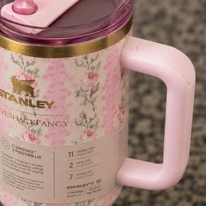 Może przedstawiać: R&oacute;żowy i kwiatowy kubek podr&oacute;żny Stanley Loveshackfancy z r&oacute;żowym uchwytem i pokrywką. Kubek posiada złoty pasek i logo Stanley. Kubek został zaprojektowany tak, aby napoje były zimne przez 11 godzin i gorące przez 7 godzin.