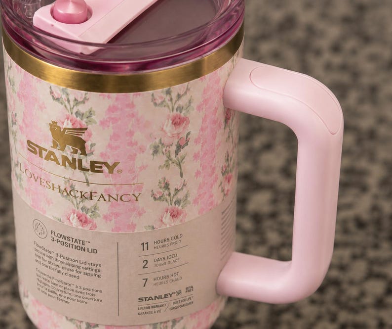 Może przedstawiać: R&oacute;żowy i kwiatowy kubek podr&oacute;żny Stanley Loveshackfancy z r&oacute;żowym uchwytem i pokrywką. Kubek posiada złoty pasek i logo Stanley. Kubek został zaprojektowany tak, aby napoje były zimne przez 11 godzin i gorące przez 7 godzin.