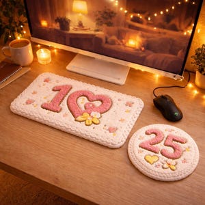 Puede incluir: Un juego de alfombrilla y posavasos de escritorio de ganchillo blanco. La alfombrilla presenta el número 10 en rosa con un diseño de corazón y detalles florales. El posavasos muestra el número 25. Ambos artículos tienen un borde festoneado.