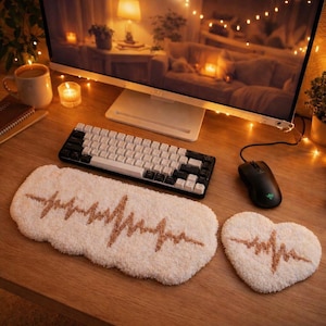 May include: A cozy desk setup featuring a white and black mechanical keyboard, a black mouse, and a cream-colored, plush desk mat with a brown heartbeat design. A heart-shaped coaster with a matching design sits nearby.