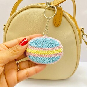 以下が含まれることがあります： パステルカラーの編み物デザインが施された、手作りの丸いキーホルダー。キーホルダーは、水色、ピンク、黄色の横縞模様が特徴です。シルバーカラーのチェーンとリングに取り付けられています。背景には、茶色のストラップが付いたベージュのハンドバッグがあります。