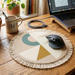 Puede incluir: Un ratón de ordenador redondo beige con un diseño geométrico en colores turquesa, mostaza y crema, bordes con flecos y un ratón inalámbrico negro. Un ordenador portátil está sobre un soporte de madera en un escritorio de madera. Se ve una taza con la palabra "Crafty".