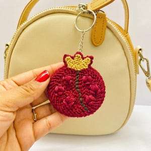 May include: A handmade red pomegranate keychain with a gold crown design. The keychain is made of embroidered fabric and attached to a silver keyring. The pomegranate has a detailed floral pattern and is held in front of a beige purse.