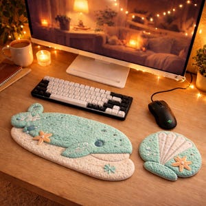 May include: A desk setup featuring a whale-shaped and seashell-shaped desk mats in shades of blue and beige. A black and white keyboard, a black mouse, and a computer monitor are also on the wooden desk.