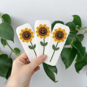 May include: Three white, rectangular crochet bookmarks, each featuring a smiling sunflower design. The sunflowers have yellow petals, brown centers, and green stems with leaves. The bookmarks are held against a backdrop of green foliage.