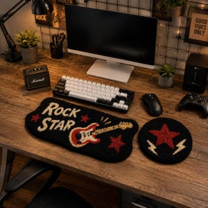 May include: A black tufted keyboard wrist rest featuring the text "ROCK STAR" in white, a red and white electric guitar graphic, and red stars. Beside it is a matching circular tufted coaster with a red star and two white lightning bolts.