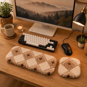 May include: A rectangular keyboard wrist rest and a square mouse pad featuring a tufted, high-pile texture with a beige and cream argyle diamond pattern. The accessories are positioned on a wooden desk surface in front of a computer setup.