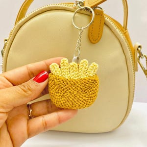 Op de afbeelding: Een handgemaakte, gele en bruine gehaakte sleutelhanger in de vorm van een bakje friet. De sleutelhanger is bevestigd aan een zilveren ketting en ring. De achtergrond omvat een beige en gele handtas.