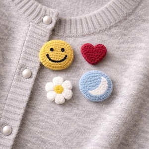 May include: A pale grey cardigan featuring four crochet patches: a yellow smiley face, a red heart, a white daisy with a yellow centre, and a blue circle with a white crescent moon. The sweater has pearl buttons.