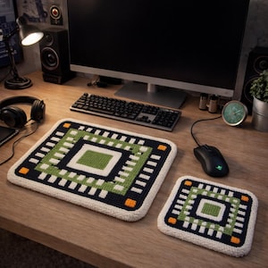May include: A square, crocheted mouse pad and coaster set. The design features a central green square, surrounded by a black square, white and green rectangles, and orange corner accents. The set is on a wooden desk.