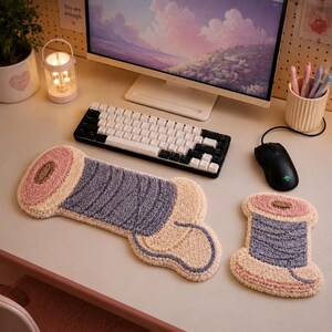 May include: A tufted desk mat and matching coaster shaped like spools of thread. The spool design features a light pink top and bottom with a textured purple thread wrap in the center. The items are placed on a desk with a keyboard and mouse.