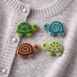 Könnte beinhalten: Vier handgefertigte Schildkröten-Anstecknadeln aus Häkelarbeit in Grün-, Blau- und Brauntönen werden auf einer grauen Strickjacke präsentiert. Die Schildkröten-Anstecknadeln haben detaillierte Schalenmuster und kleine schwarze Augen.