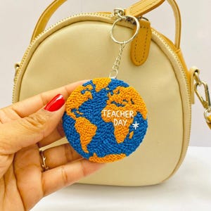 Könnte beinhalten: Ein Schlüsselanhänger zum Lehrertag mit einem Globus-Design. Die Ozeane sind blau, die Kontinente orange und der Text "Teacher Day" ist weiß gestickt. Der Schlüsselanhänger ist rund und an einer silbernen Kette befestigt.