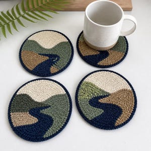 May include: Four round coasters with a landscape design. Each coaster features a river winding through hills in shades of green, tan, and cream, with a dark blue border. A white mug rests on one coaster. The coasters are made of a textured material.
