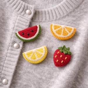 Puede incluir: Primer plano de un cárdigan gris adornado con cuatro parches de frutas de ganchillo: una rodaja de sandía, una rodaja de naranja, una rodaja de limón y una fresa. El cárdigan tiene tres botones blancos.