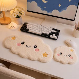 May include: A cream-colored, cloud-shaped keyboard wrist rest and a matching smaller mouse pad, both featuring embroidered black eyes, a smiling mouth, pink cheeks, and yellow stars. The textured, plush fabric surfaces are soft and cushioned.