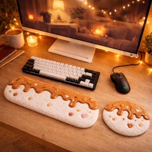 Puede incluir: Una configuración de escritorio con un teclado mecánico en blanco y negro, un ratón negro y un reposamuñecas largo, blanco y marrón con detalles de corazón. También hay un posavasos redondo a juego. El escritorio tiene un monitor y una taza de café.