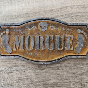 Puede incluir: Un letrero metálico rectangular con un acabado oxidado y desgastado que presenta la palabra "MORGUE" en letras grises en relieve. El letrero incluye un pequeño icono de calavera sobre el texto y dos huellas de pies a ambos lados.